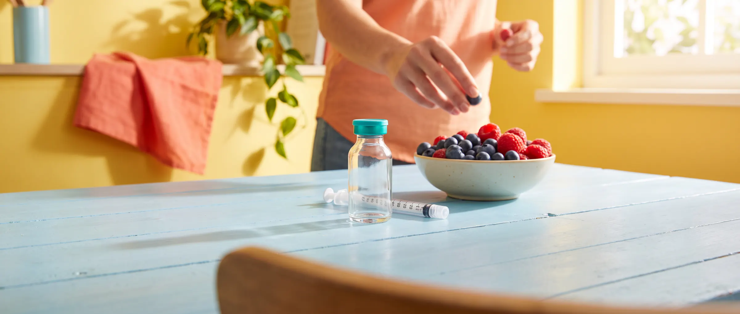 A plain vial beside a capped syringe on a pastel-blue table with berries